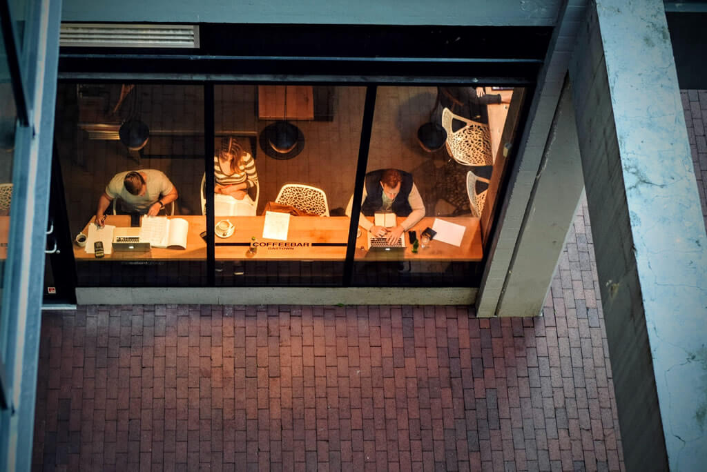 People working on laptops behind a large window, seen from above